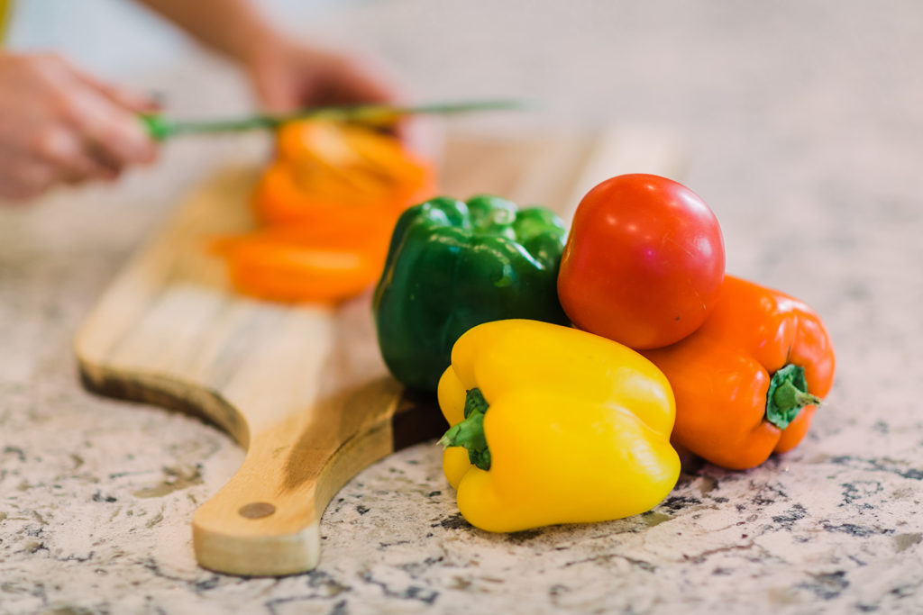 Green, yellow, and orange peppers and a red tomato in the foreground. Jen Lyman, dietitian at New Leaf Nutrition, cuts an orange pepper on a wooden cutting board in the background