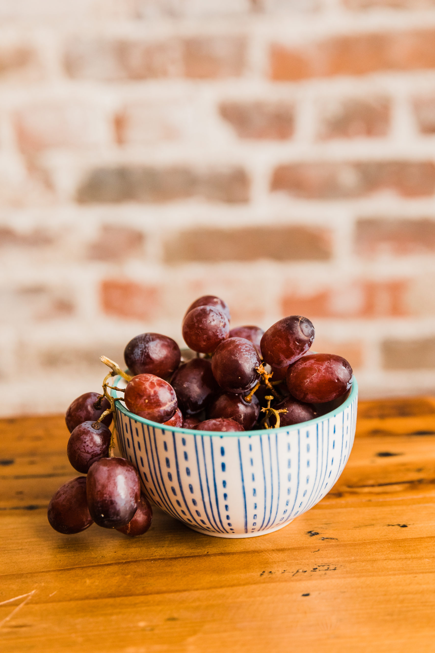 Red grapes cascading out of a small patterned bowl onto a wooden table in front of a brick wall backdrop